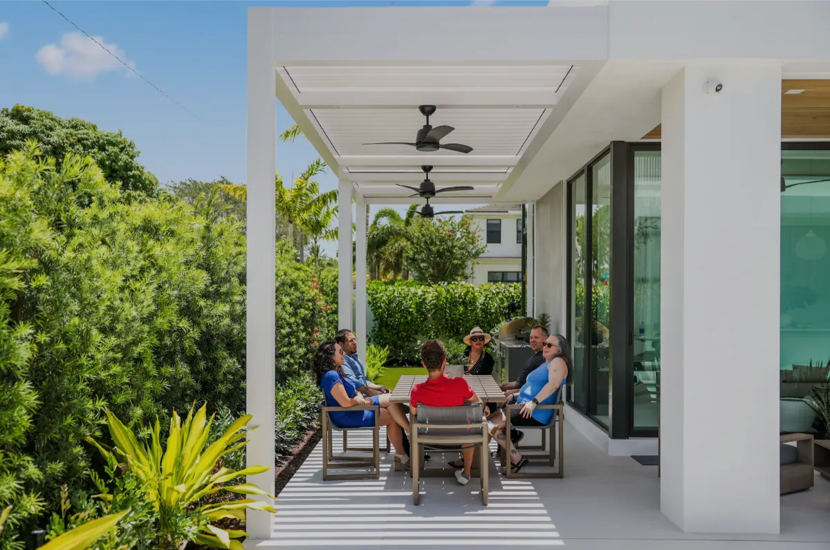 Group enjoying a shaded patio beneath a smart pergola with ceiling fans, adjacent to a modern glass-walled home.