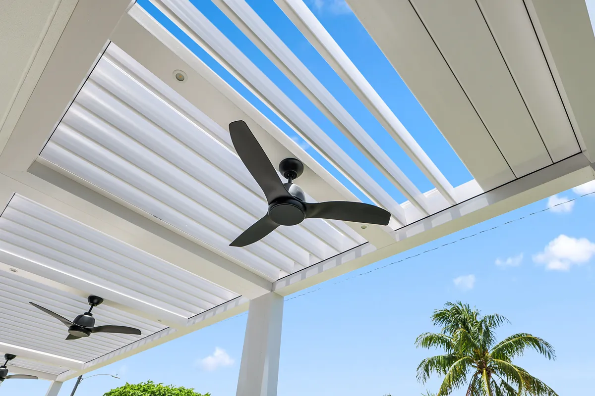 Modern white pergola with motorized louvers and ceiling fans, showcasing airflow and sun control against a clear blue sky.