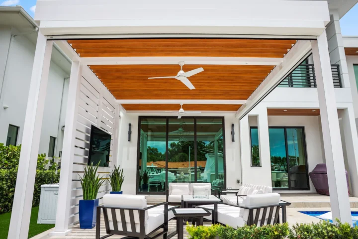 Modern white R-Blade pergola with wood slat ceiling and ceiling fans covering outdoor patio seating area next to contemporary home with large glass doors