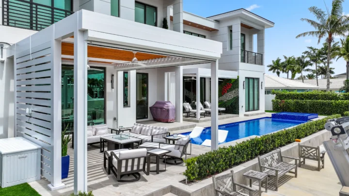 Modern white R-Blade pergola with wood slat ceiling and ceiling fans covering outdoor patio seating area next to contemporary home with large glass doors