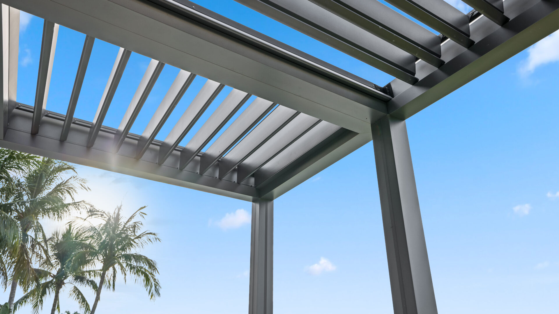 Close-up of aluminum louvered roof on L-shaped pergola with palm trees and blue sky in background.