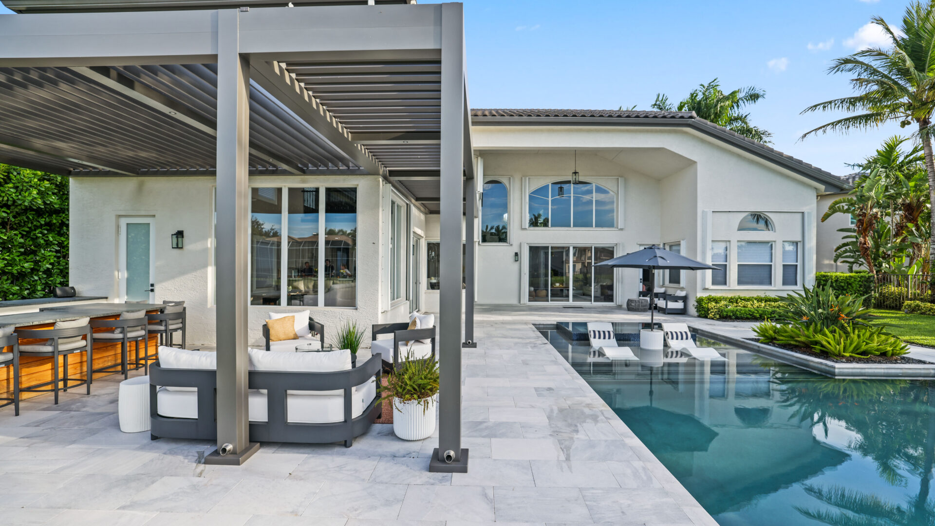 Modern L-shaped pergola with outdoor kitchen and lounge area overlooking luxury pool and landscaped yard.