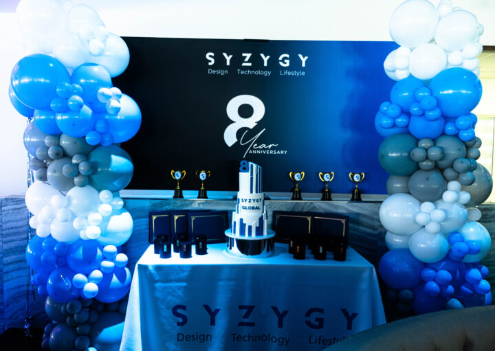 SYZYGY Global 8th anniversary cake, trophies, and awards display with blue and white balloon décor.