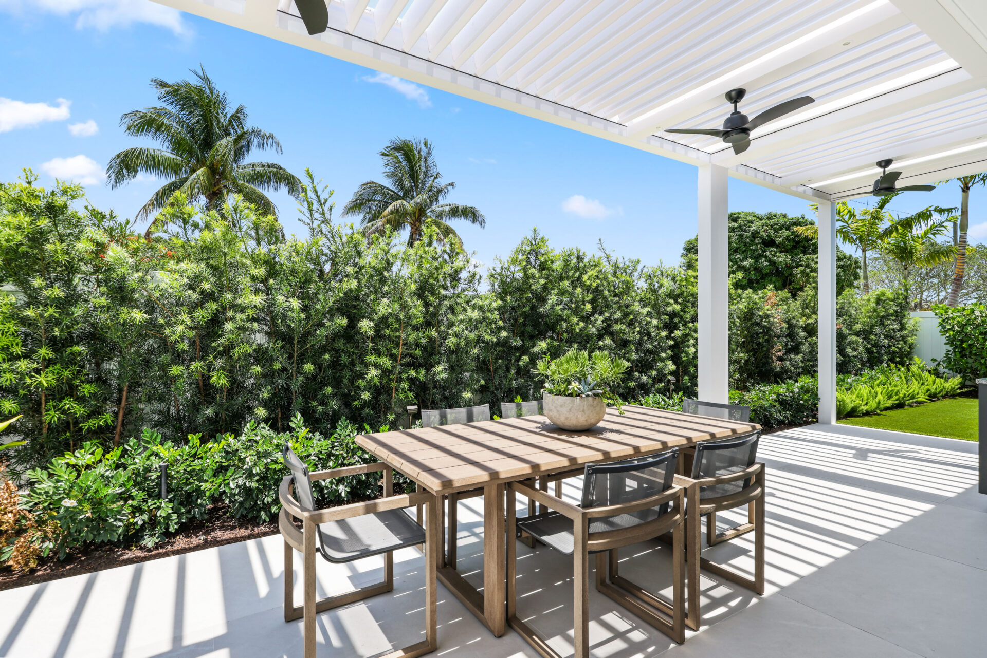 White L-shaped pergola with louvered roof shading modern outdoor dining area surrounded by tropical greenery.