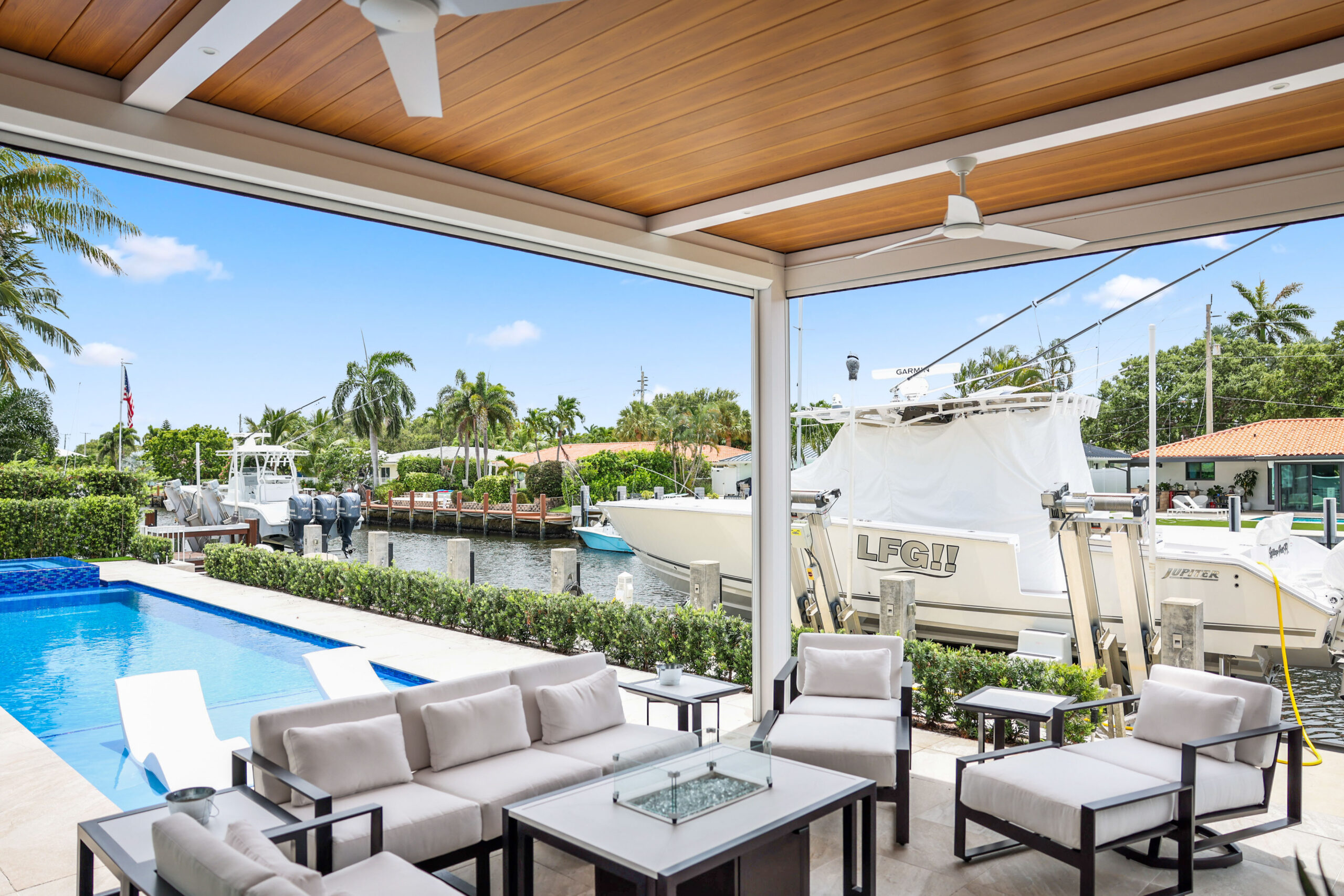Wood grain aluminum patio cover shades a waterfront seating area with pool and boat dock views.