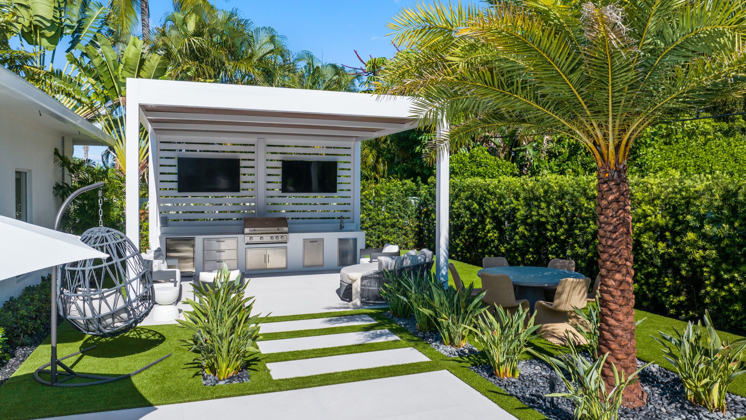 Modern outdoor kitchen under a sloped pergola roof with TV, seating, and tropical landscaping in South Florida.