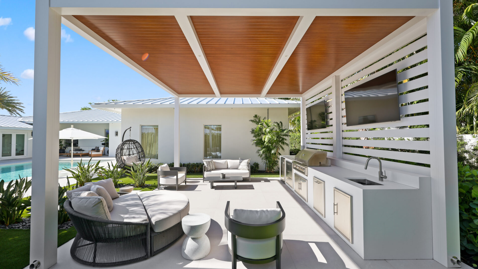 Luxury outdoor kitchen with sloped pergola roof, wood ceiling panels, seating, and poolside lounge area.