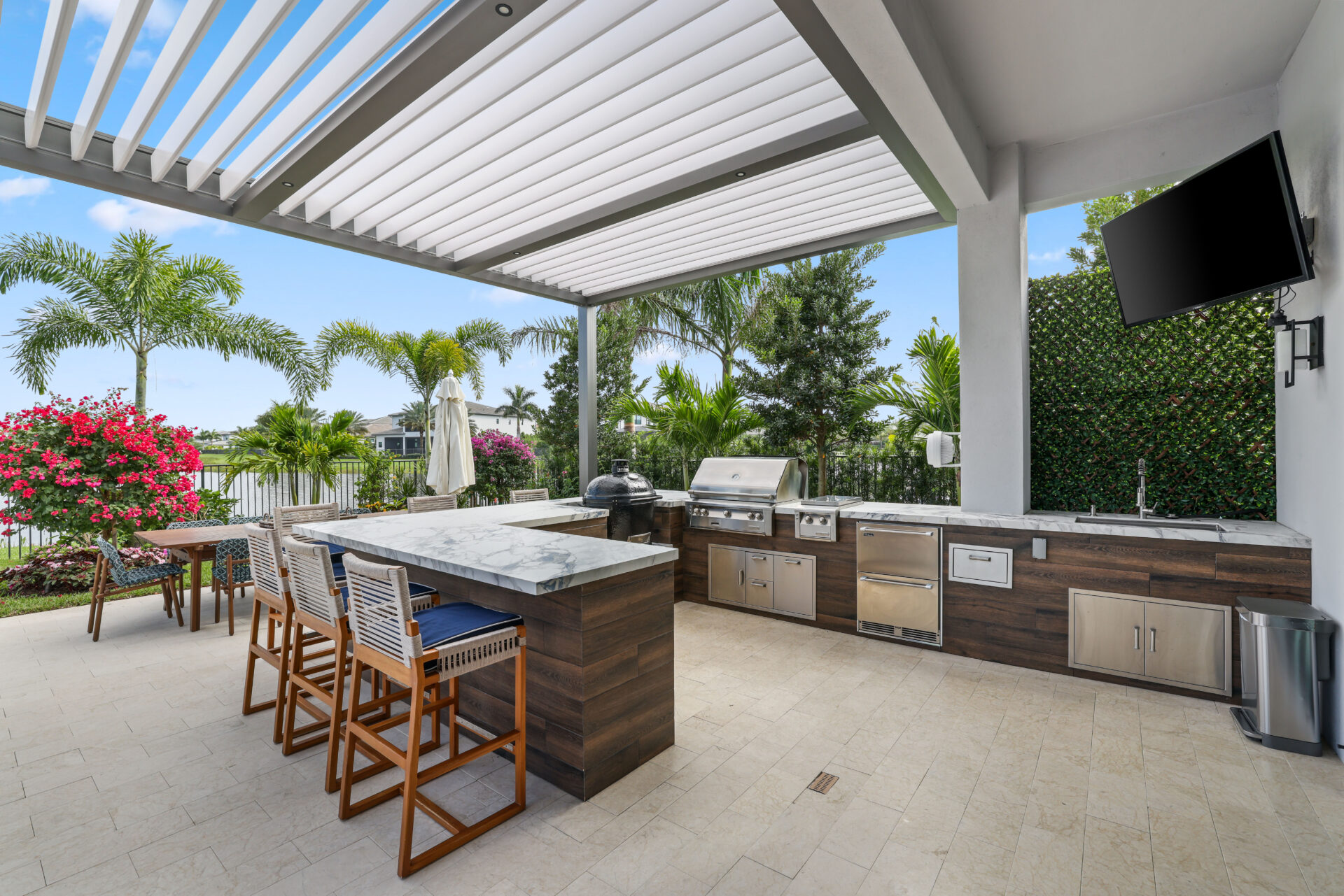 Modern outdoor kitchen with marble counters, built-in grill, and R-Blade pergola overlooking lush landscaping.