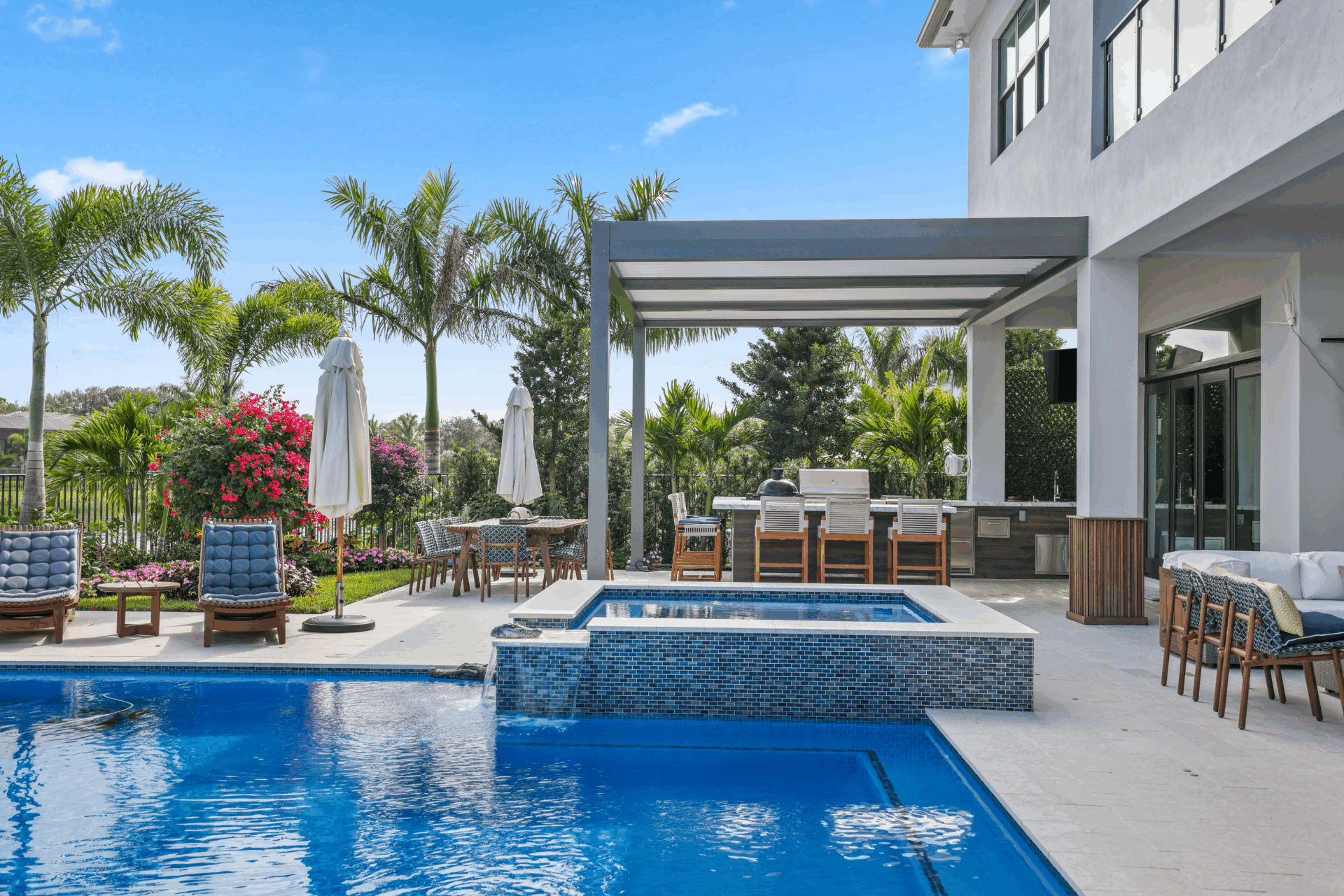 Luxury poolside outdoor kitchen with R-Blade pergola, bar seating, and lush tropical landscaping in Boca Raton.