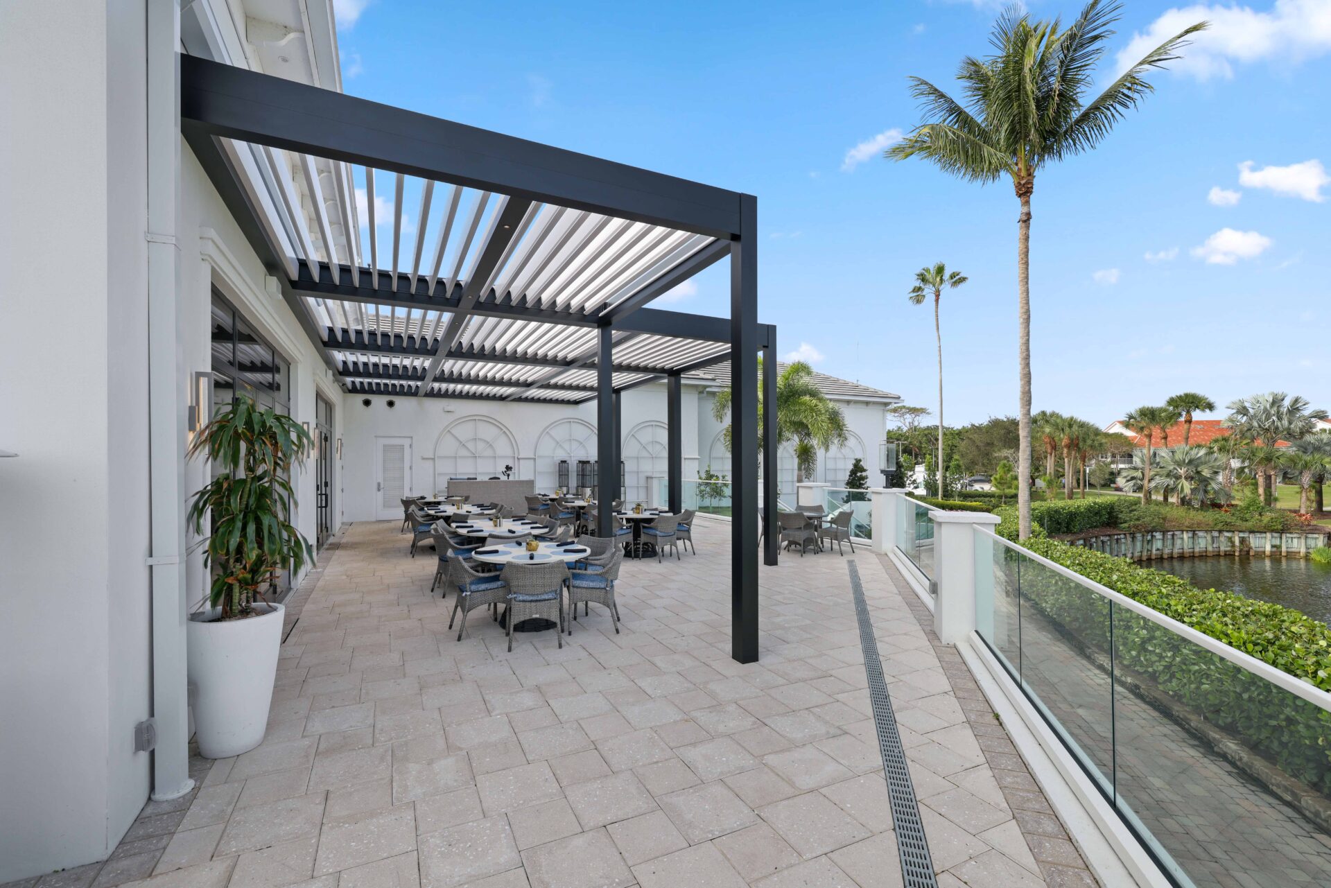 Outdoor dining terrace at Broken Sound Club with R-Blade pergolas and glass railing overlooking lush landscaping and water.