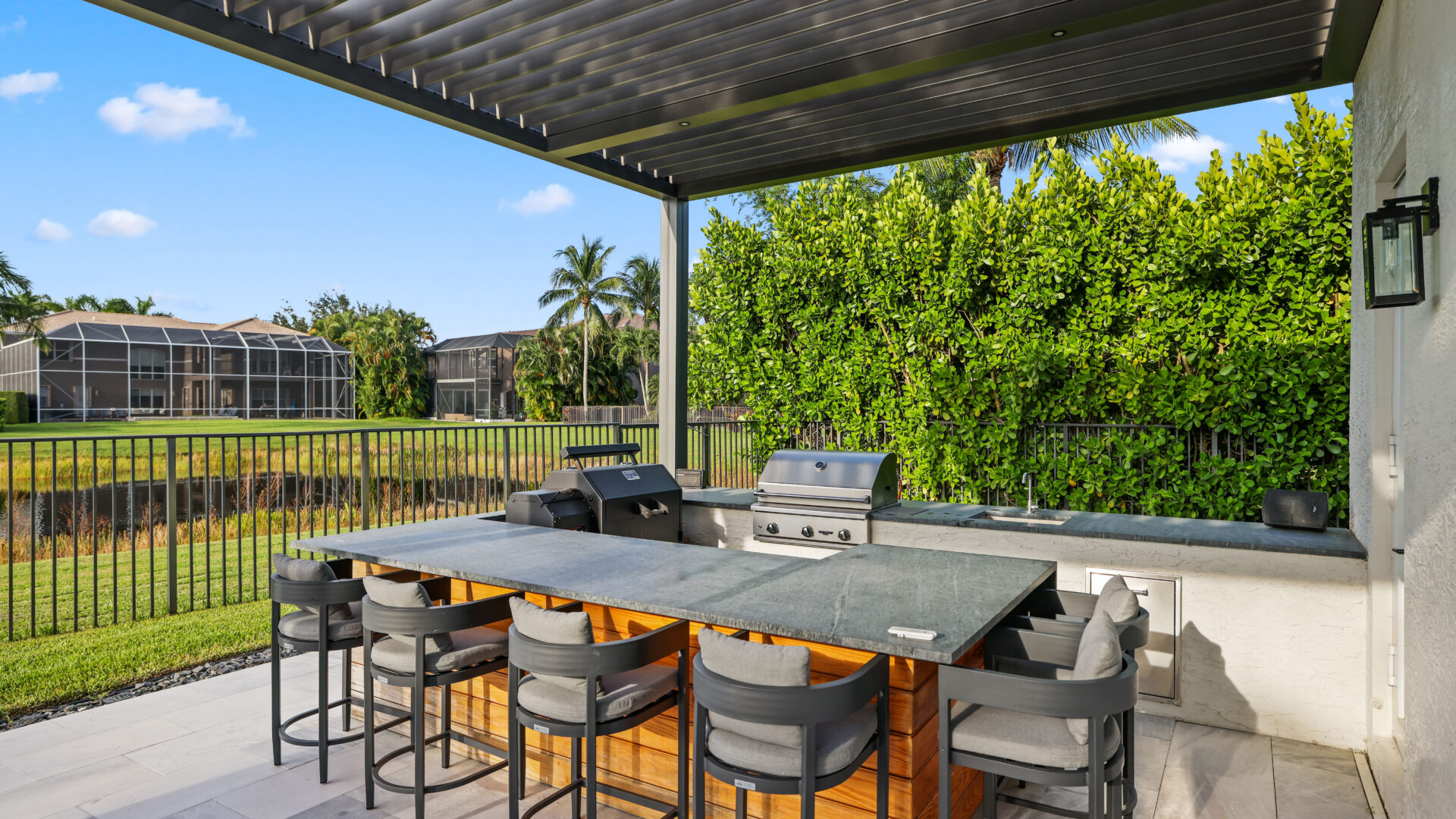 Louvered pergola covering outdoor kitchen island with grill, bar seating, and open backyard views.