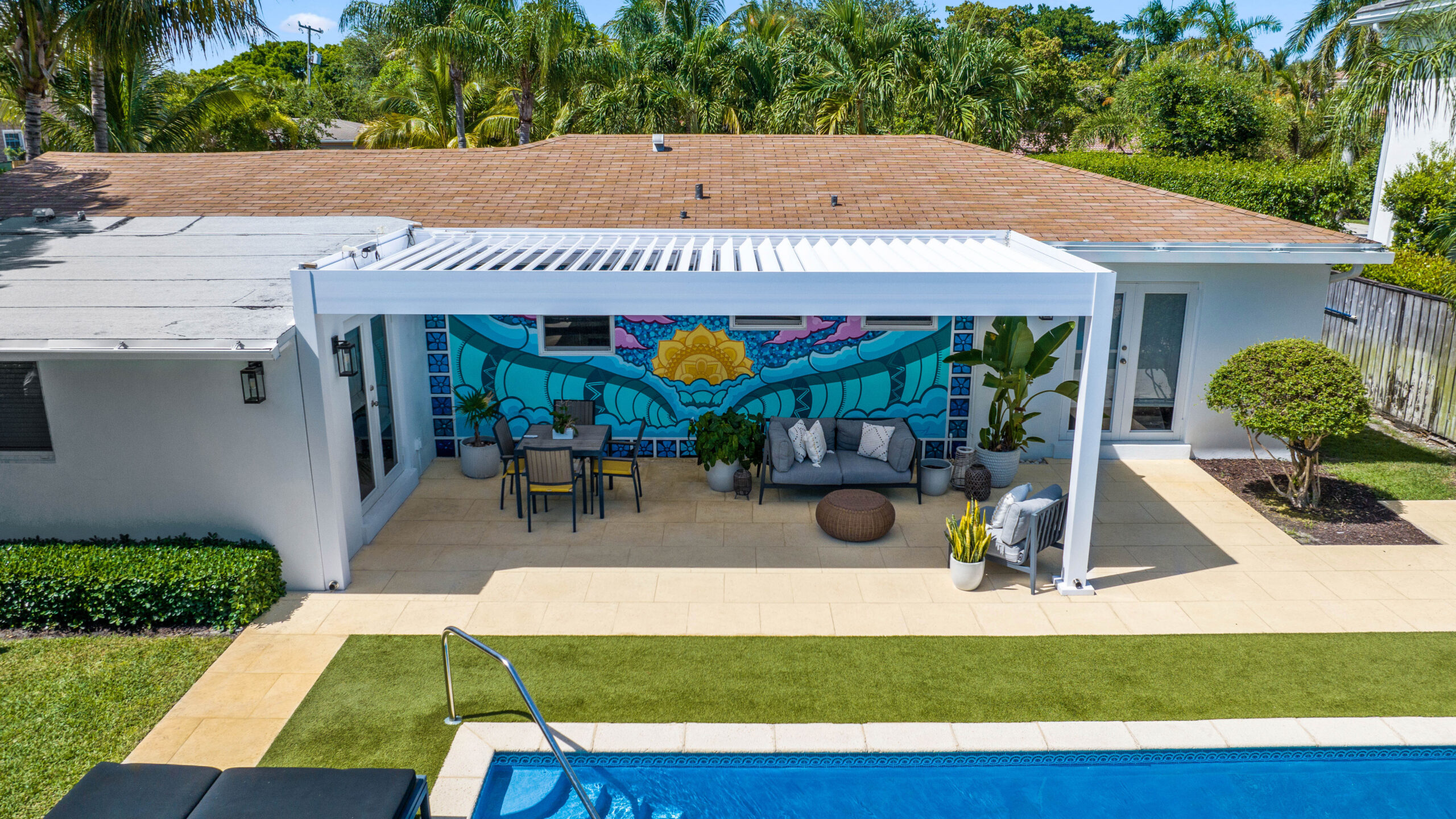 White louvered pergola over backyard patio with poolside seating and colorful mural backdrop.