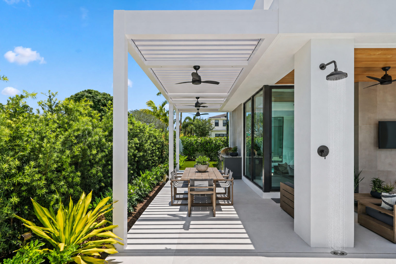 Wall-mounted outdoor shower near a modern pergola, pool patio, and lush backyard landscaping.