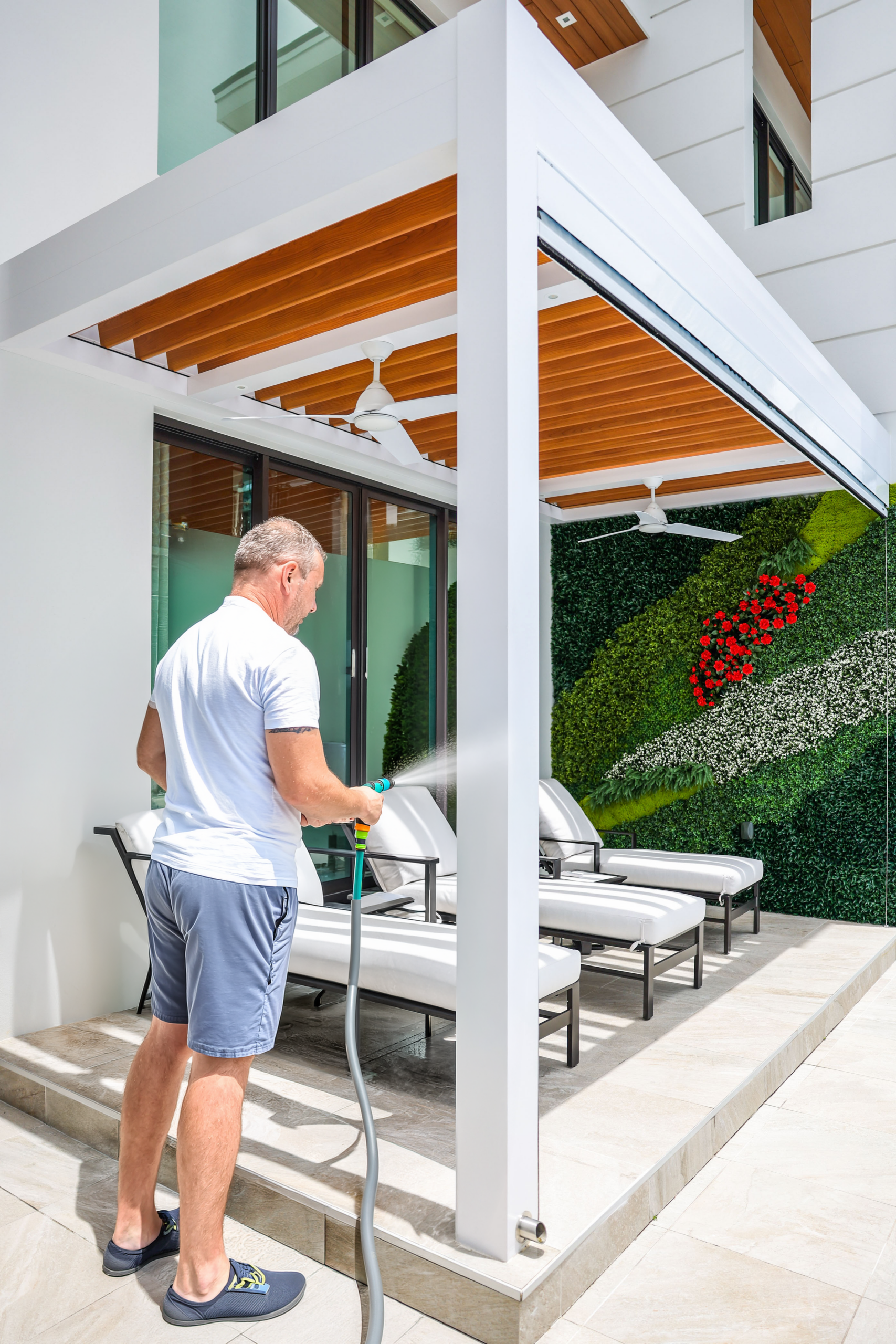 Man rinsing post of a white pergola with wood-grain louvers and living plant wall.