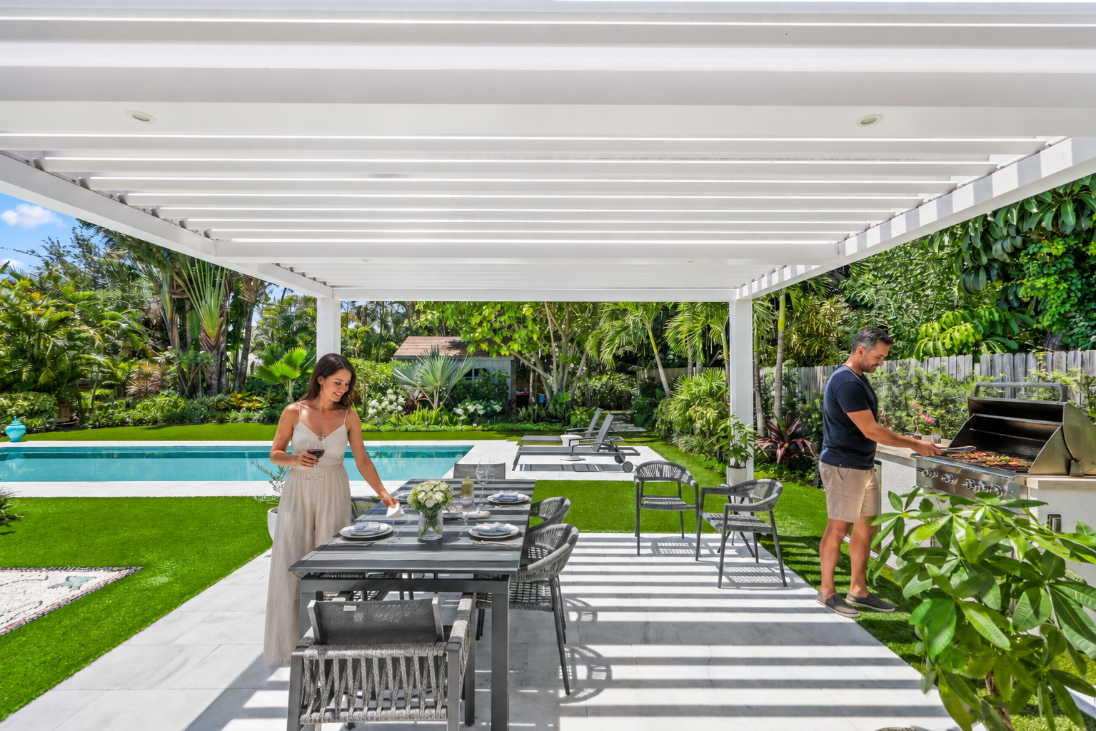 Modern backyard grill station beside pool and dining area under white louvered pergola.